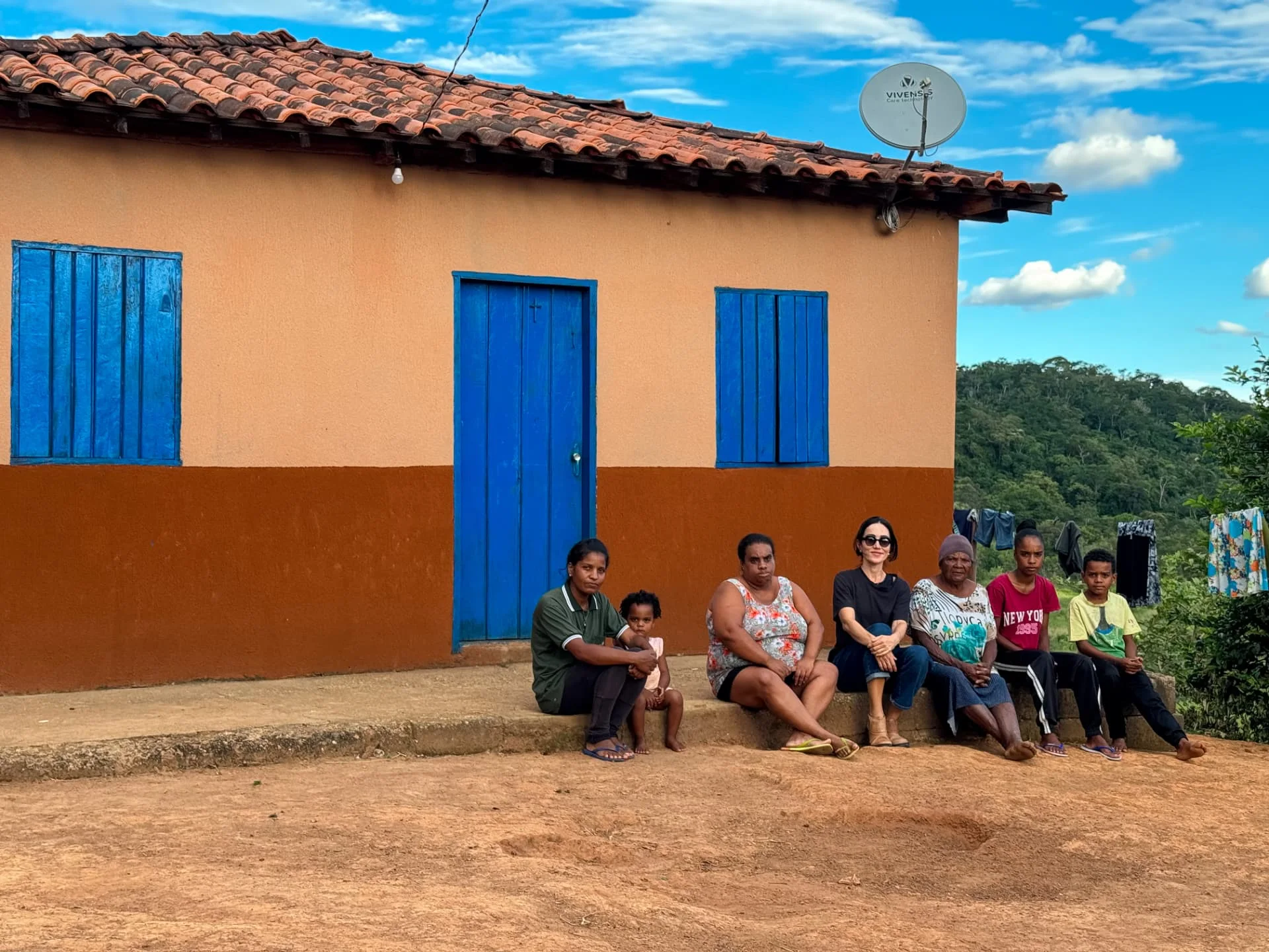 Moradoras e moradores sentados na varanda de uma casa pintada na Vila Mumbuca, com a serra ao fundo